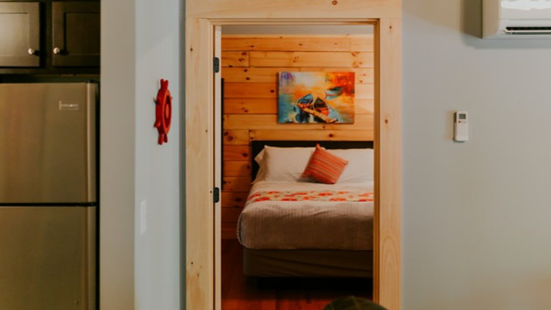 Bedroom doorway view: bed with orange pillow and artwork; wooden walls.