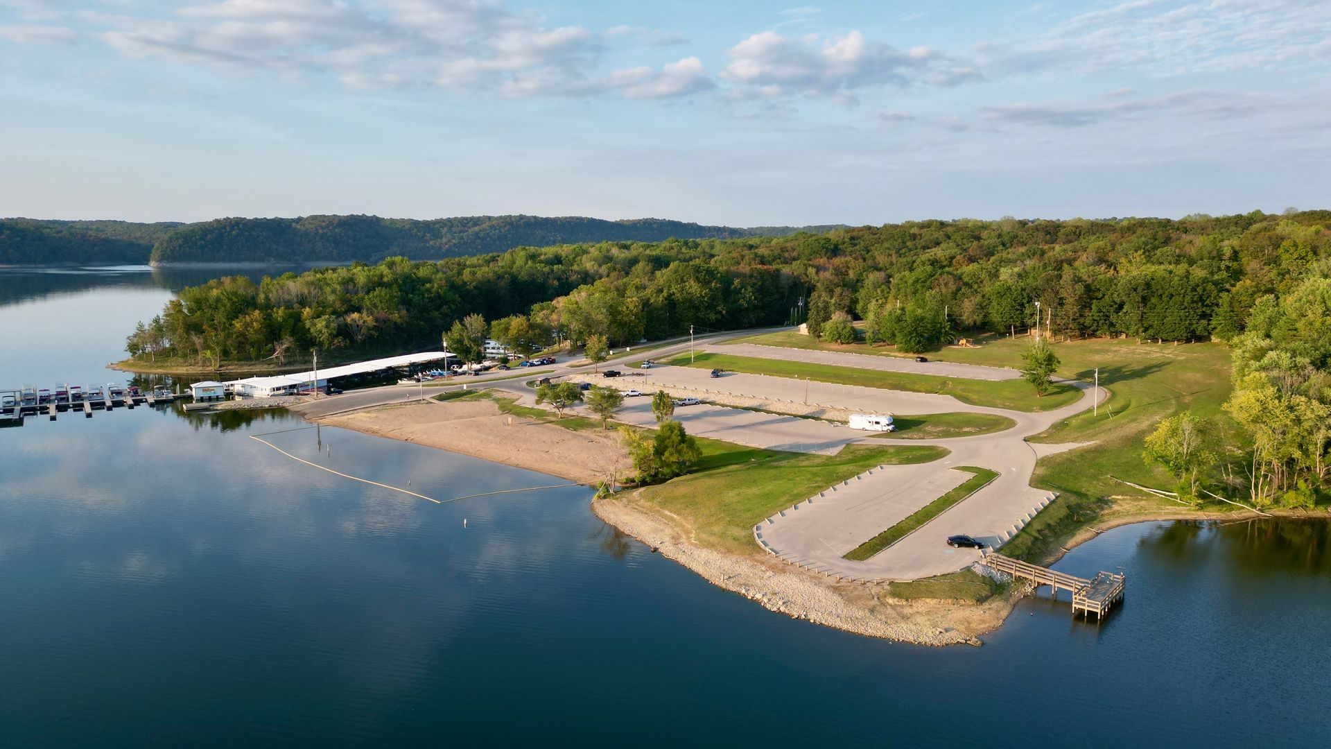 Aerial view of a lakefront park with a sandy beach, boat dock, parking, and forested hills under a blue sky.