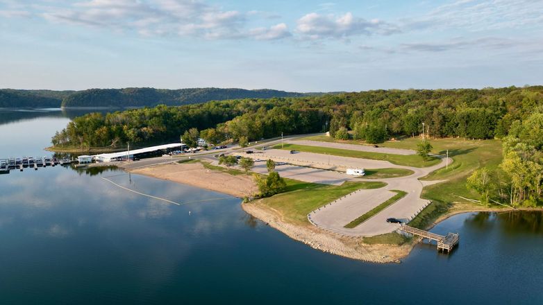 Aerial view of a lakefront park with a sandy beach, boat dock, parking, and forested hills under a blue sky.