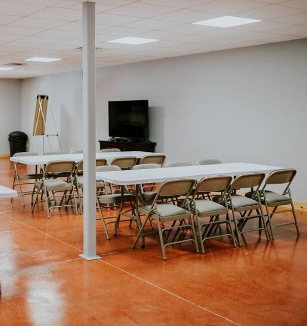 A room with tables and folding chairs, a TV, and a support beam. Orange floor, white walls.