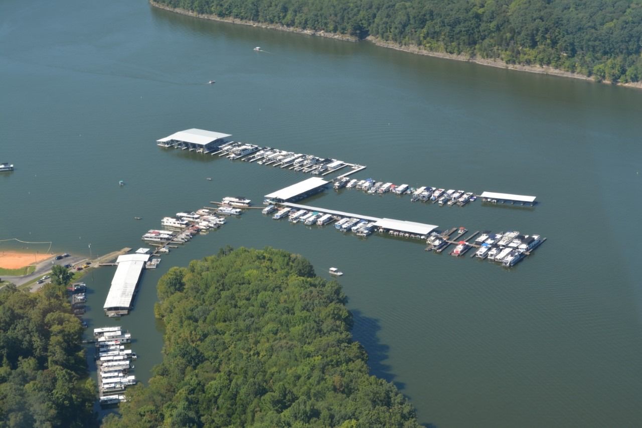 Aerial view of a marina on a lake with numerous boats docked, surrounded by green trees and water.