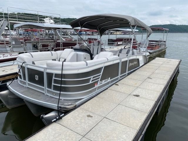 Pontoon boat docked at a pier, gray and white with a canopy, lake in background.