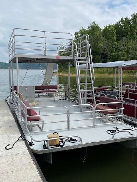 Double-decker pontoon boat docked on a lake, featuring stairs, railings, seating, and a second-story deck.