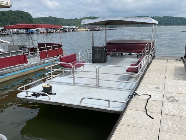 Pontoon boat with burgundy seats docked at a pier on a lake. Overcast sky and other boats in the background.