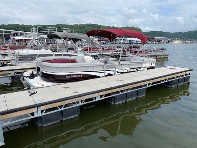 Pontoon boat with burgundy awning docked at a marina. Water, other boats, and hills in the background.