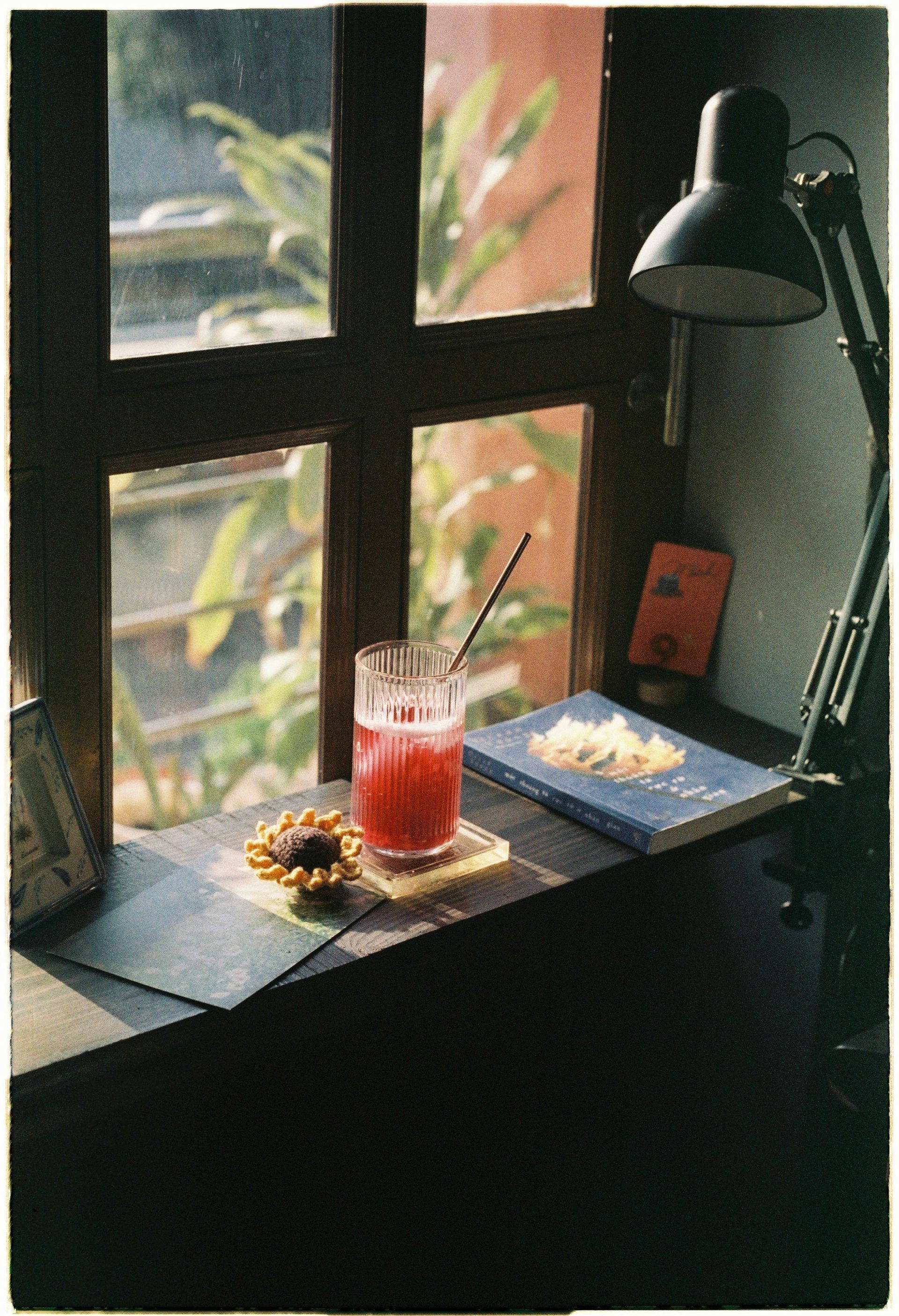 A glass of red liquid is sitting on a table next to a book and a lamp.