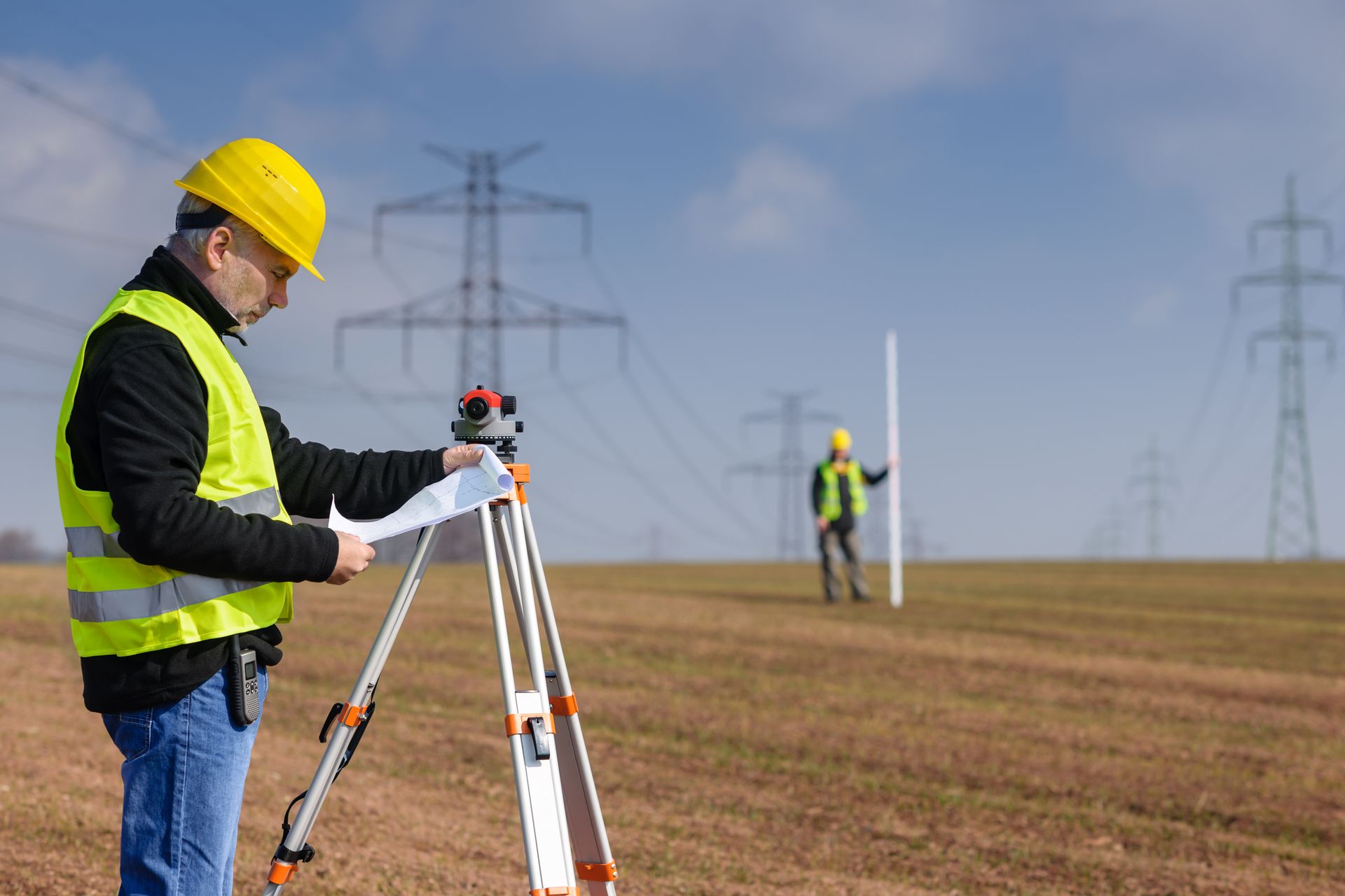 A man in a yellow hard hat is standing in a field with a tripod.