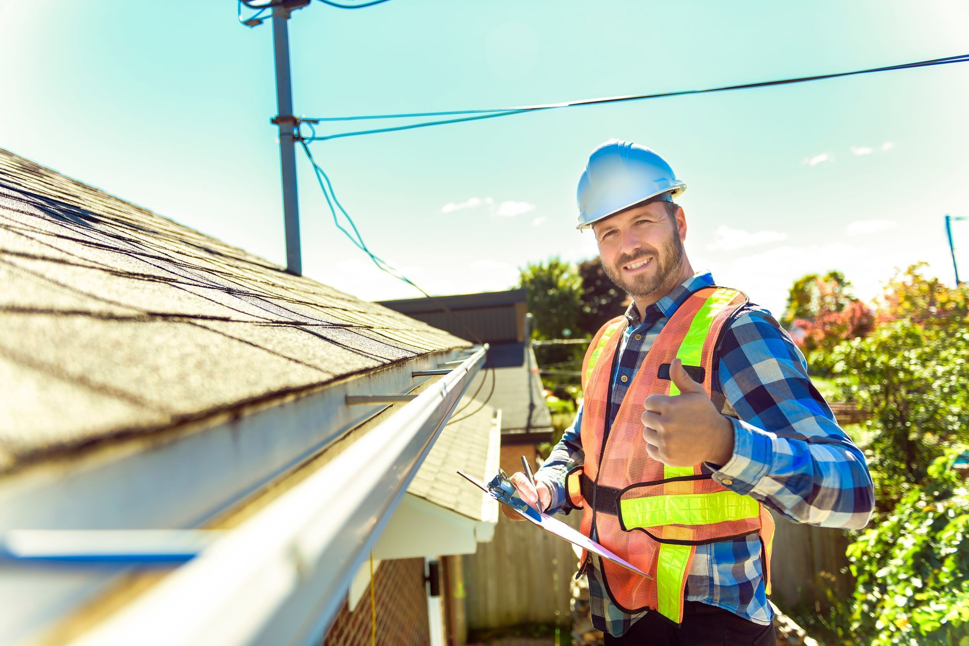 A man is standing on top of a roof holding a clipboard and giving a thumbs up.