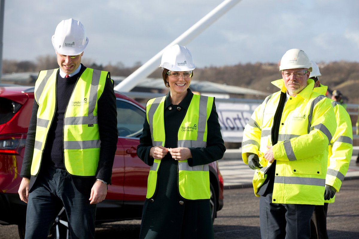 Three people wearing hard hats and yellow vests are walking in front of a red car.