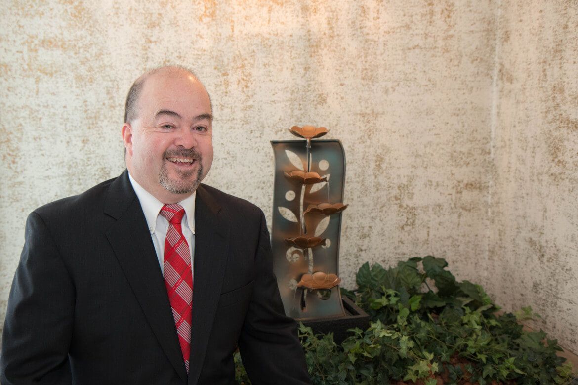 Man in suit and red tie smiles near a decorative water fountain and greenery.