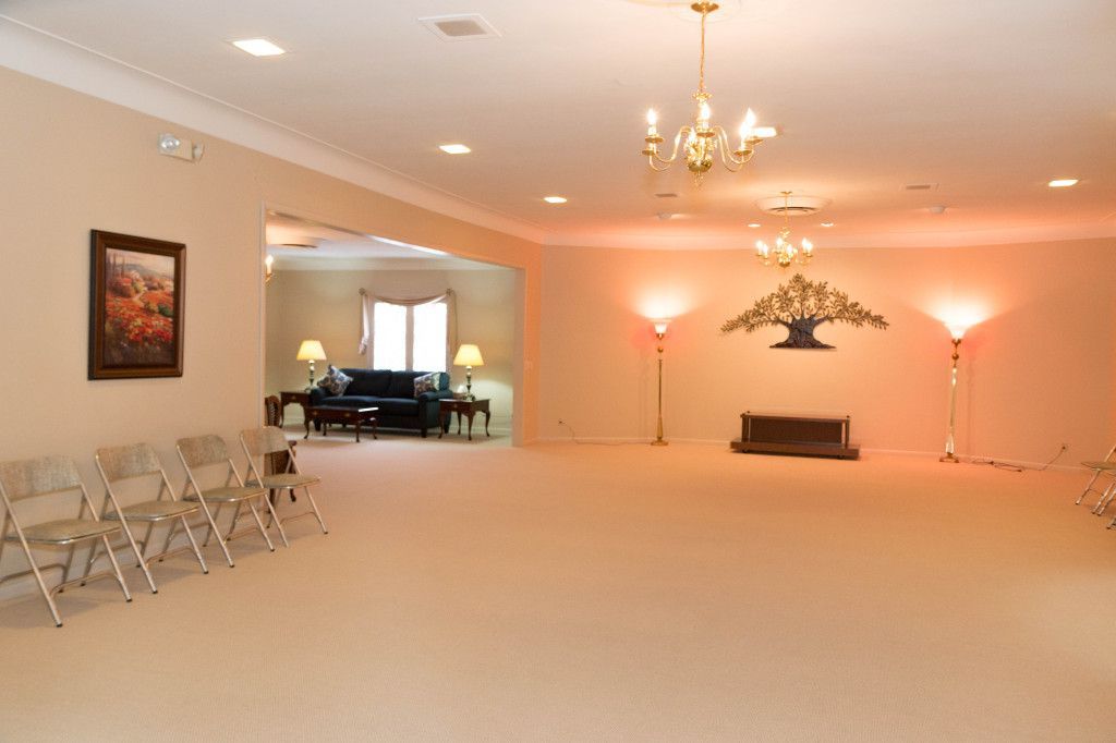 Empty beige-carpeted room with chandelier and several rows of chairs, leading into a secondary room with furniture.