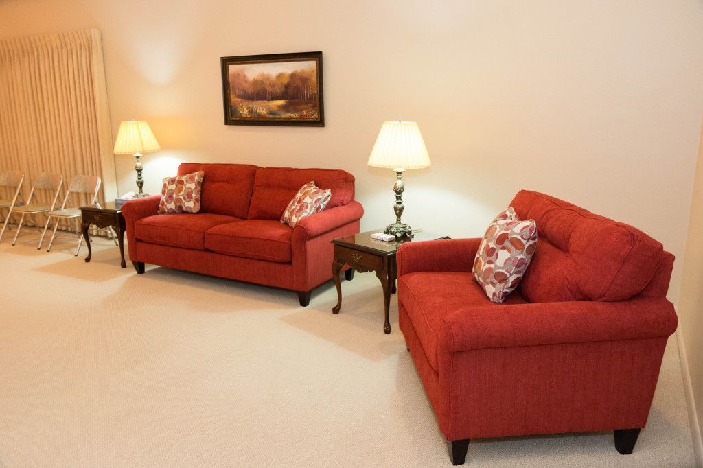 Red couch and armchair with floral pillows, lamps, and a painting in a living room.