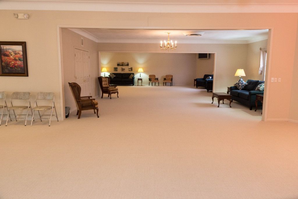 Large, beige-carpeted room with seating areas and an open doorway. Several chairs and a sofa are visible.