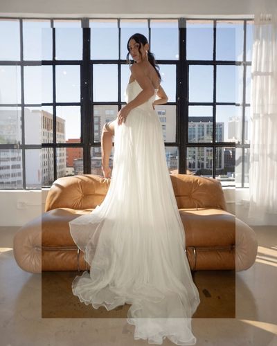 A woman in a wedding dress is standing next to a couch in front of a window.