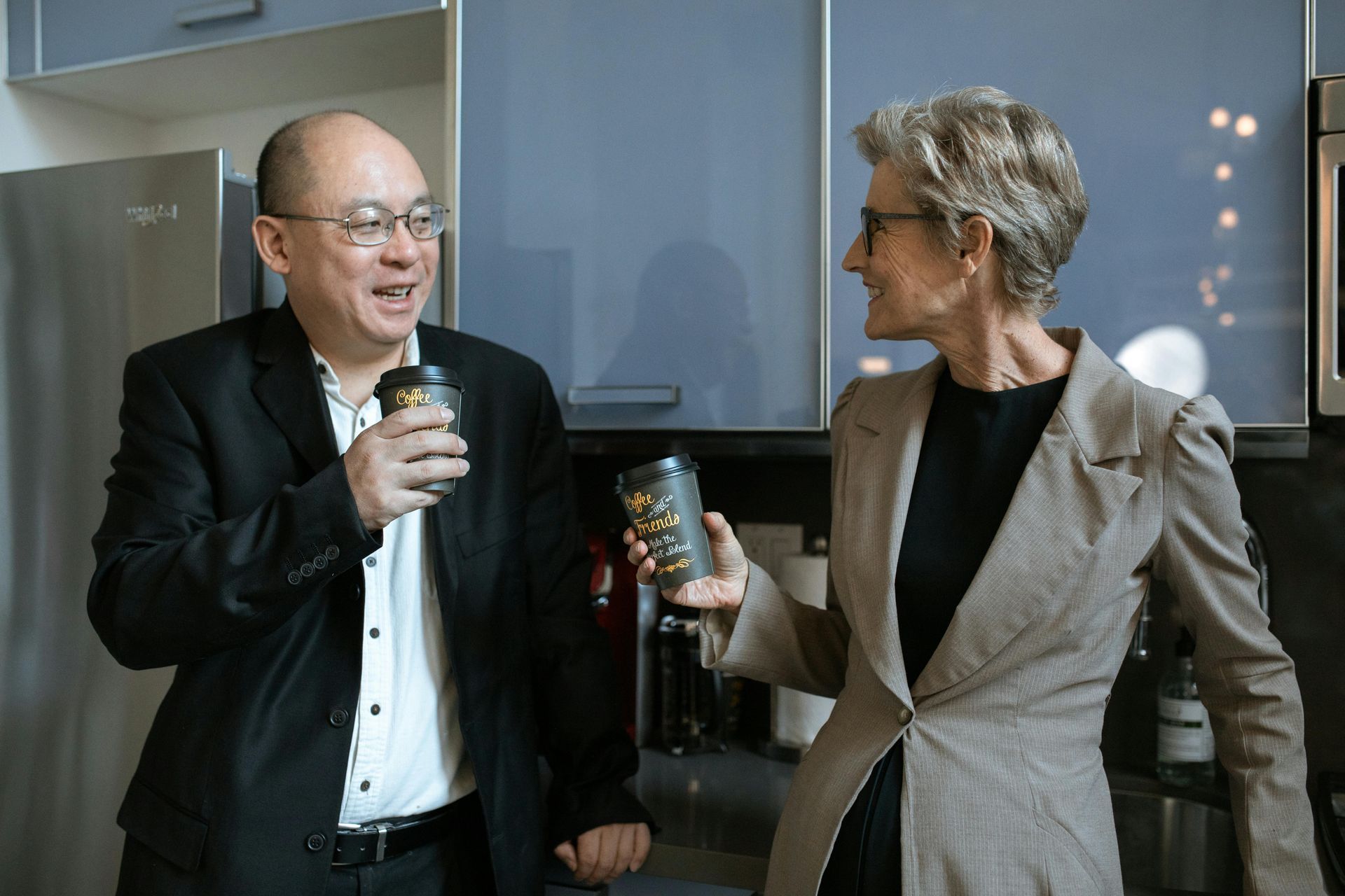 A man and a woman are standing in a kitchen drinking coffee.