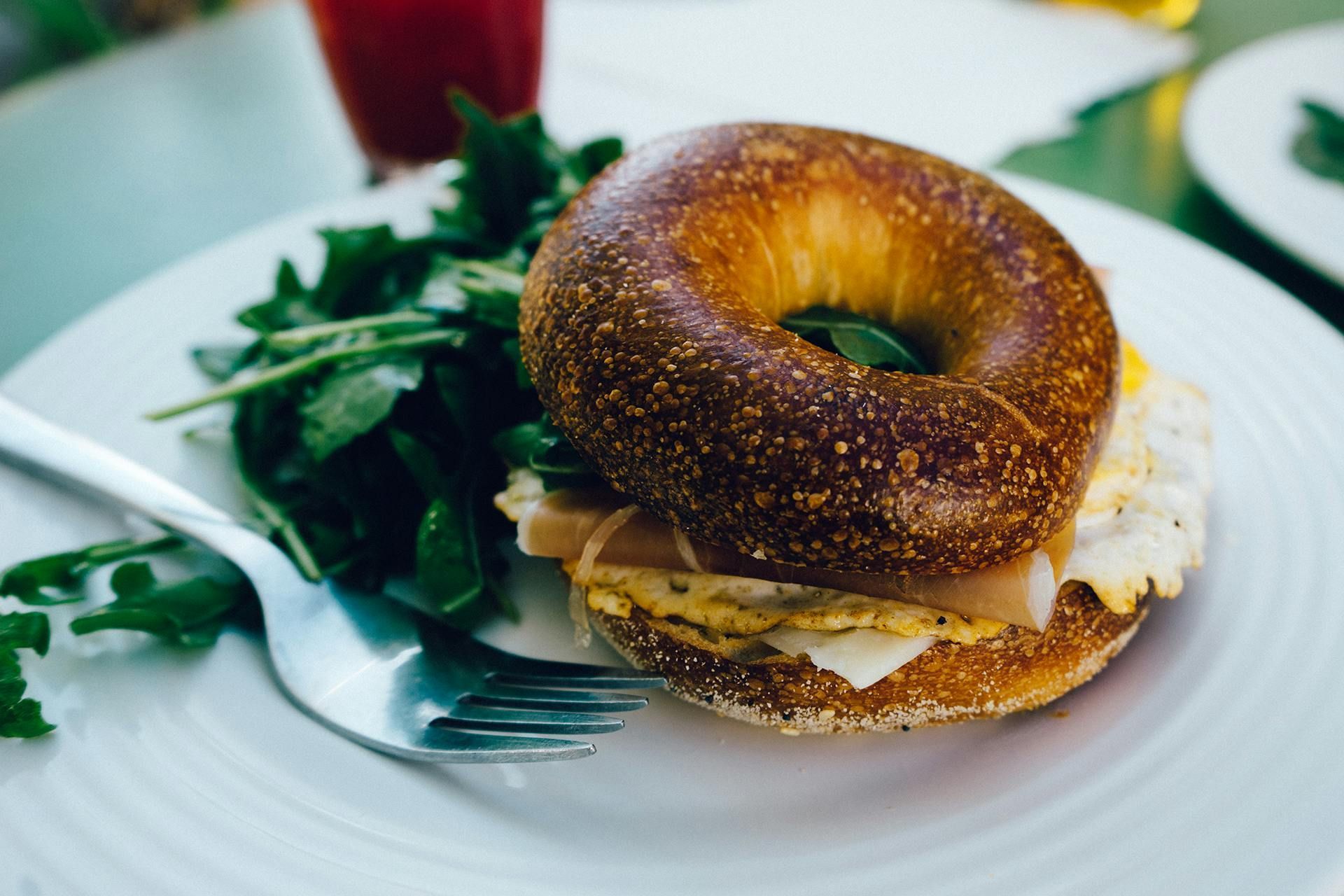 Bagel sandwich with egg and ham on a white plate with arugula and a fork.