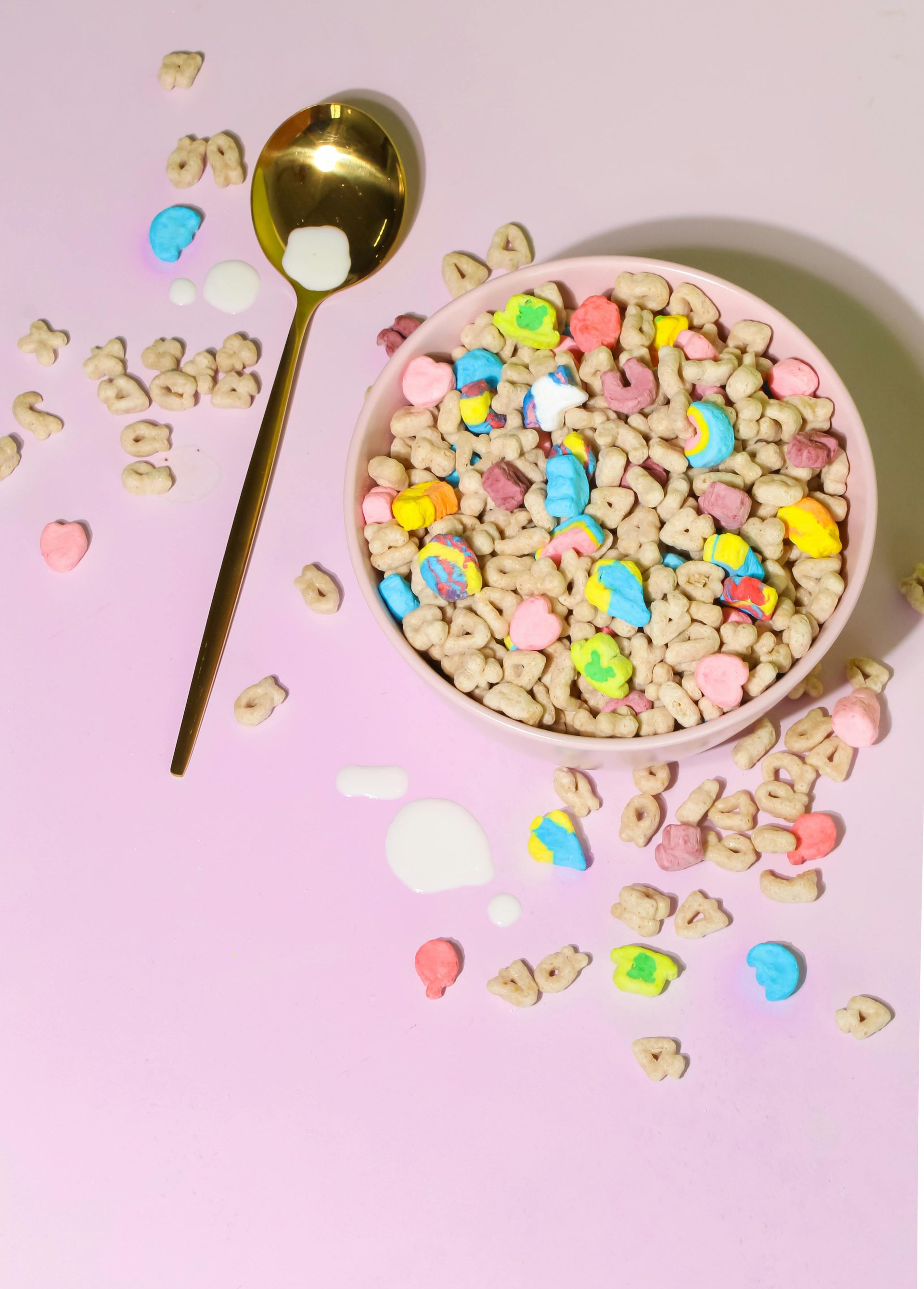 A bowl of Lucky Charms cereal with a golden spoon on a pink background.