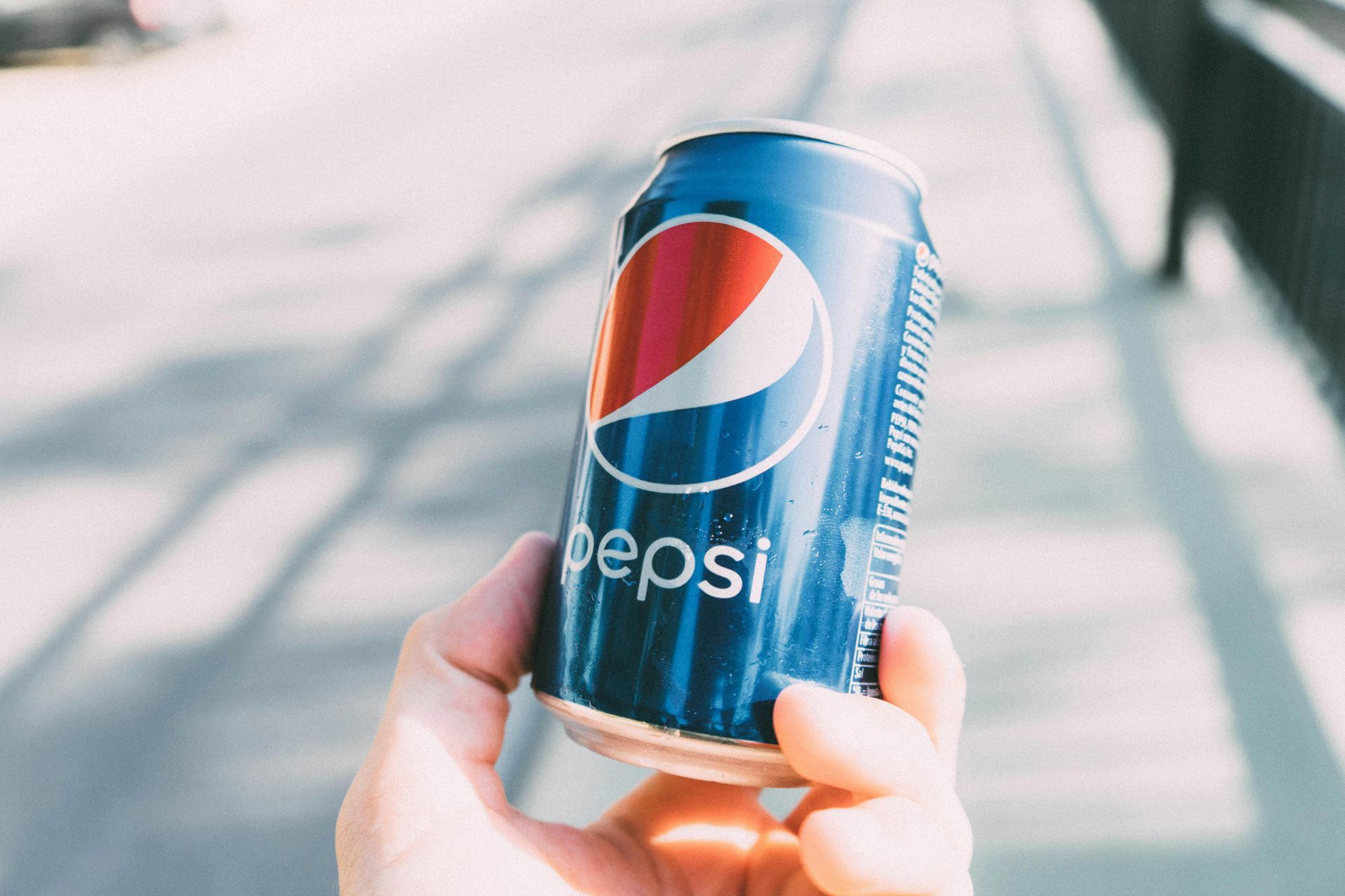 Hand holding a Pepsi can, blue and red branding, outdoors in daylight.