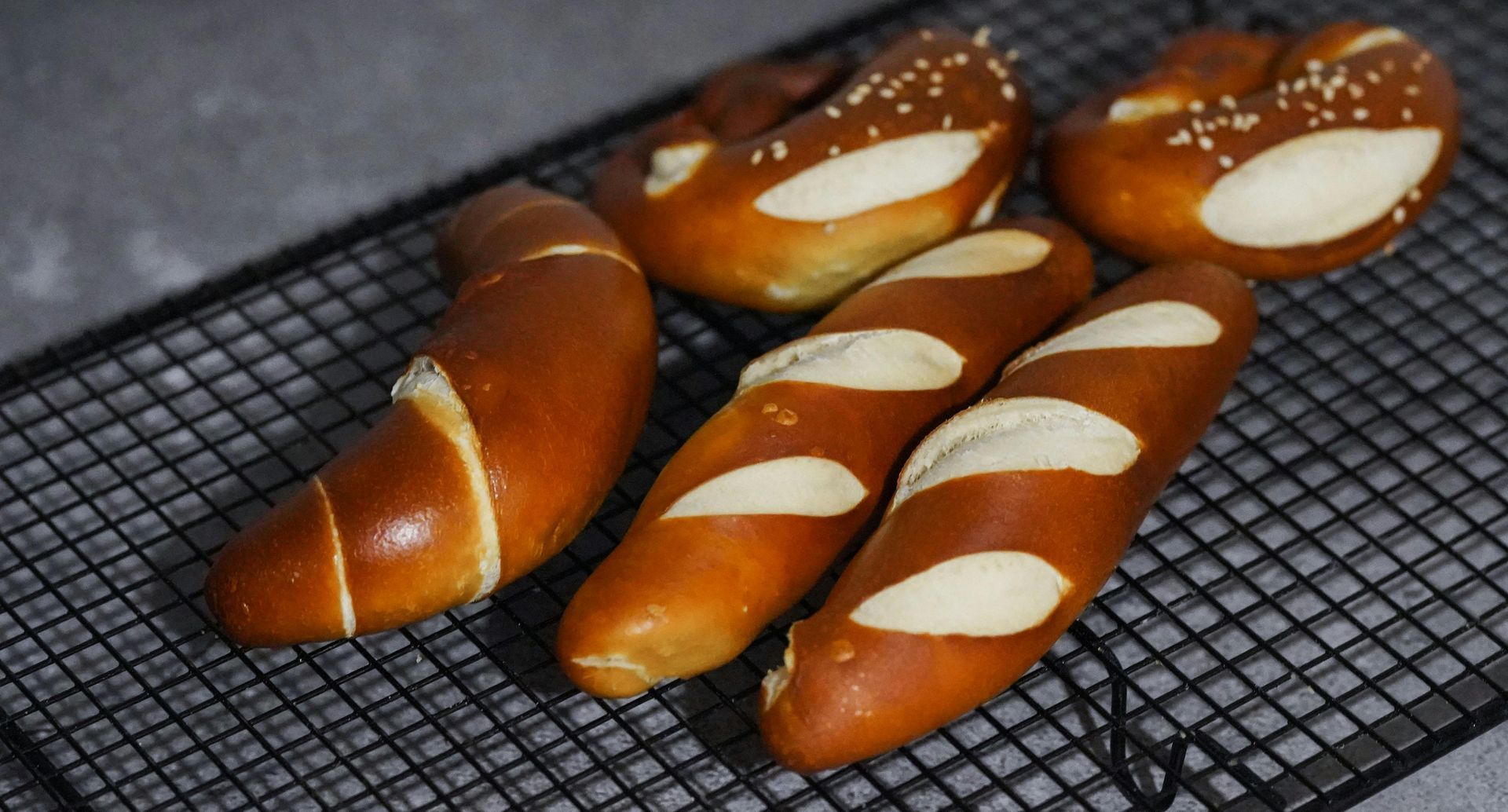 Pretzels and pretzel bread rolls on a black wire rack, against a gray background.