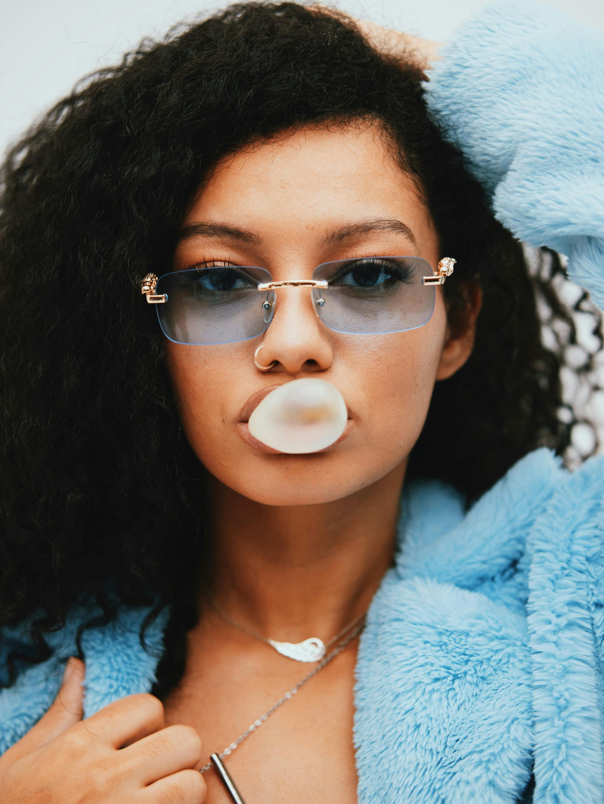 Woman blowing bubble gum, wearing blue-tinted glasses and fluffy blue robe.