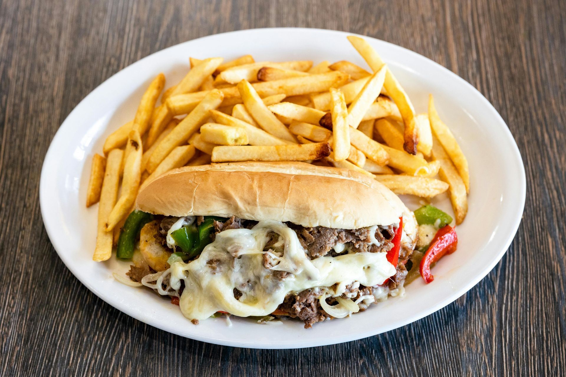 Cheesesteak sandwich with peppers, onions, and fries on a white plate.