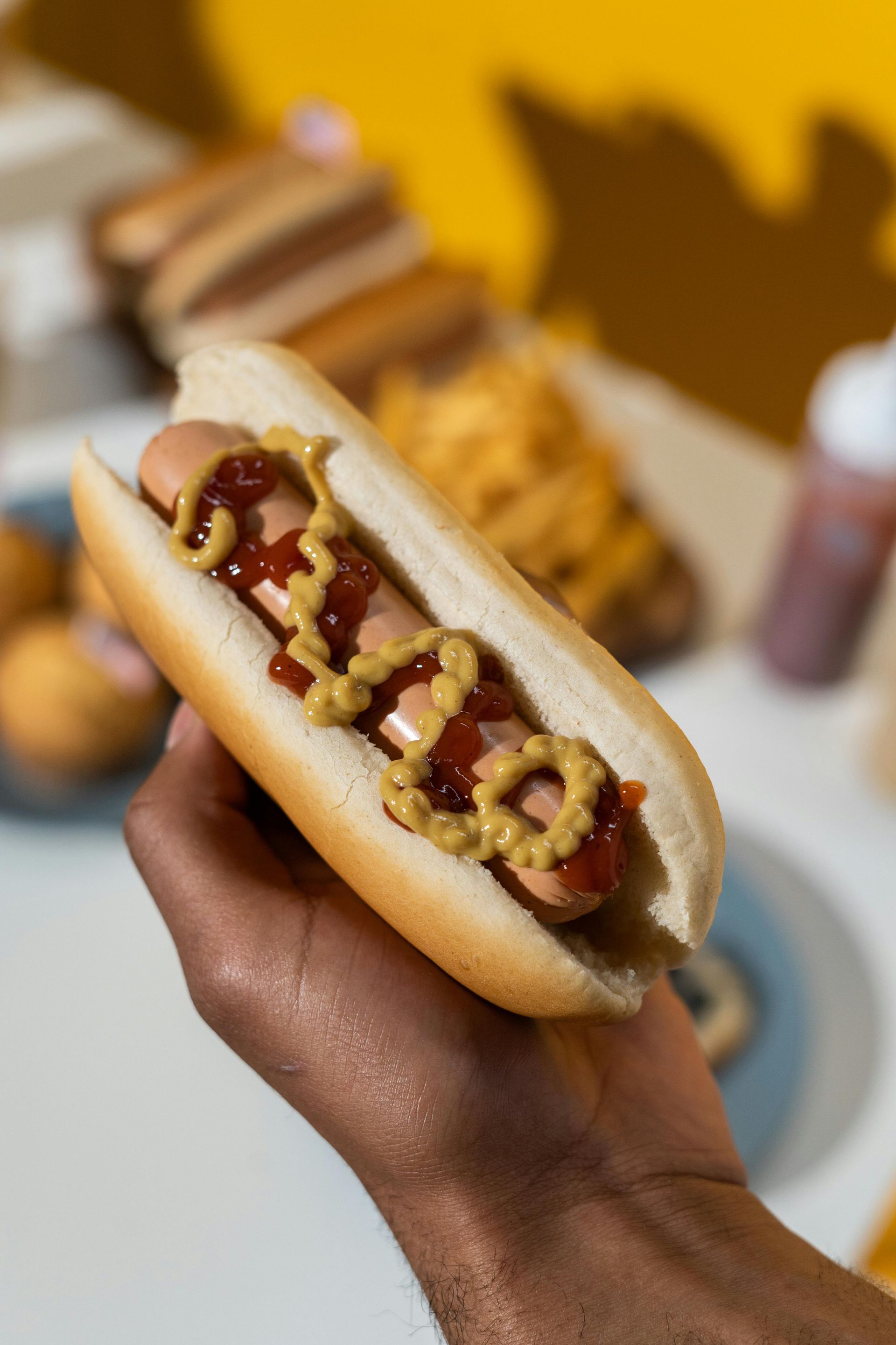 Hand holding a hot dog with ketchup and mustard on a bun. Other hot dogs and food are in the background.