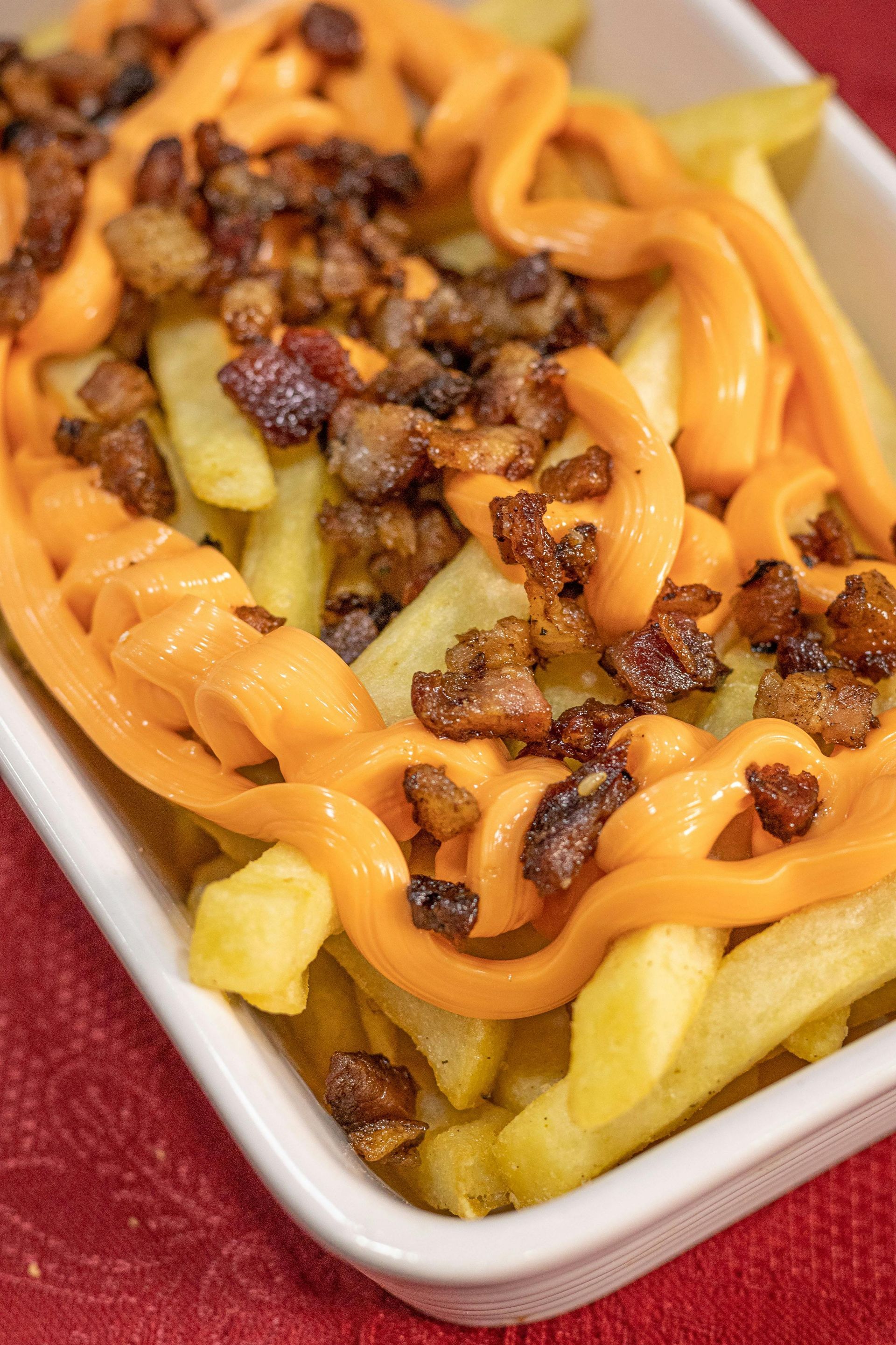 Fries in white dish, topped with orange cheese sauce and chopped bacon.