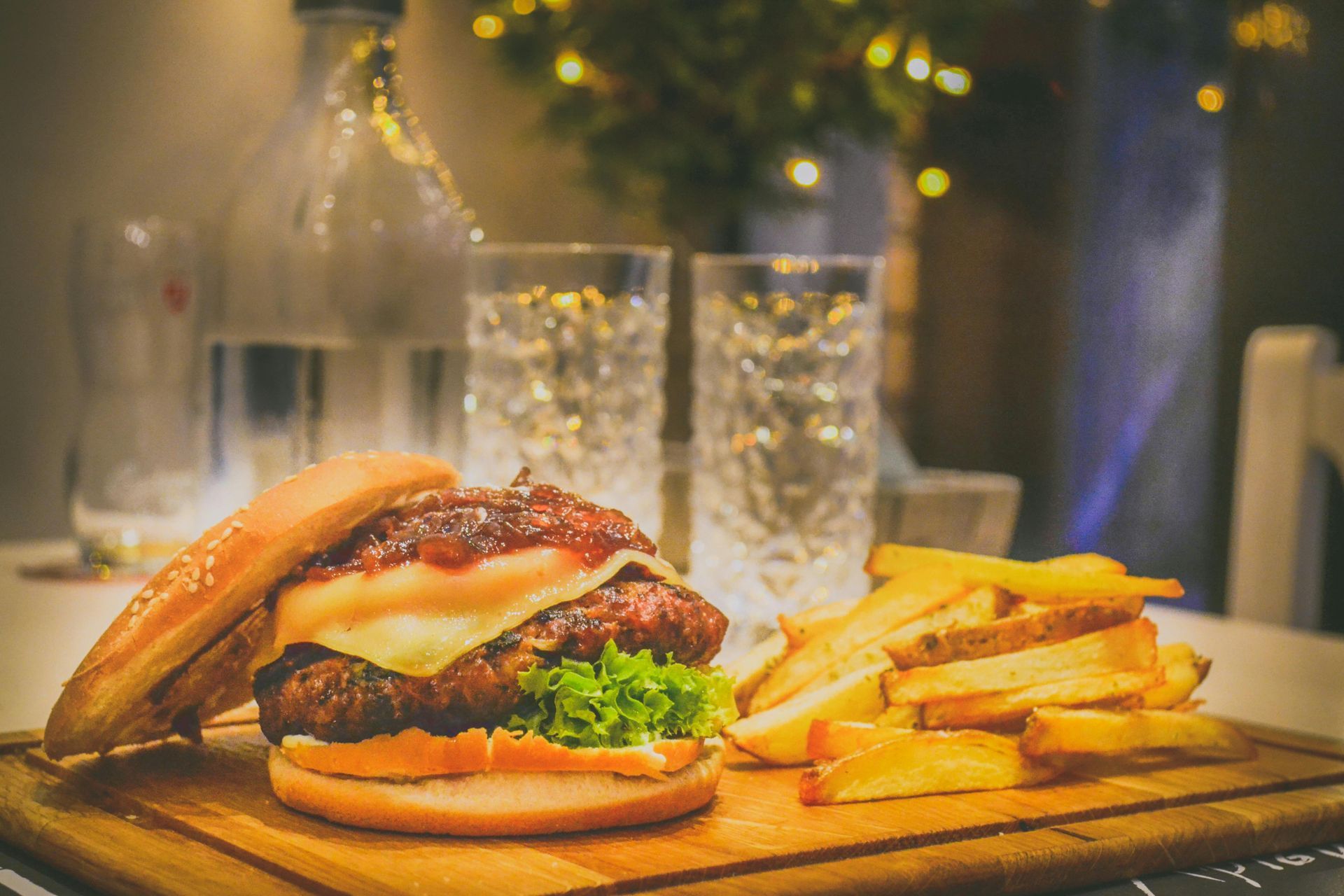 Burger with fries on a wooden board, with glasses and bottle in the background, dimly lit setting.