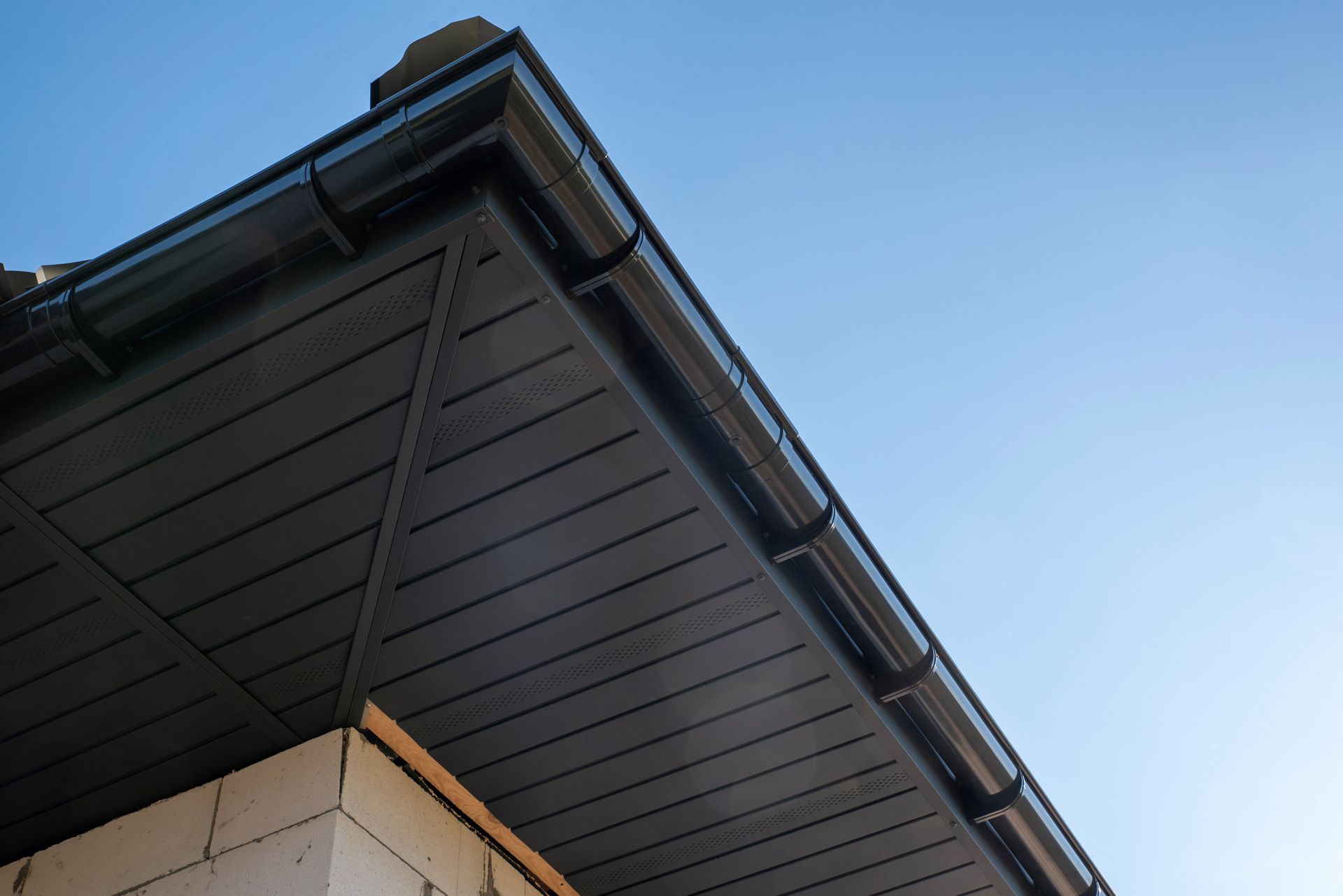 Dark brown wooden soffits and eaves with black gutters installed on the corner of a house against a clear blue sky.
