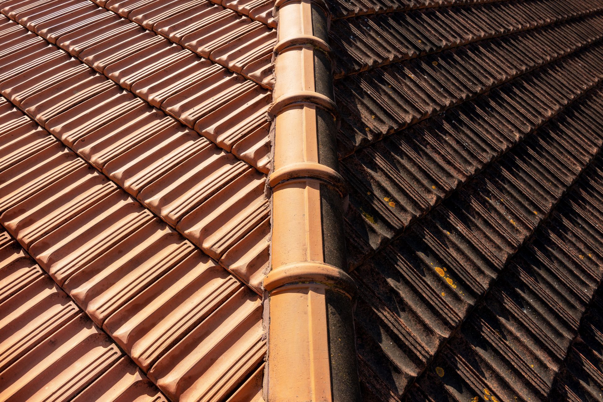 A high-angle view shows a roof ridge separating a clean, light-brown tiled section from a dark, weathered tiled section.