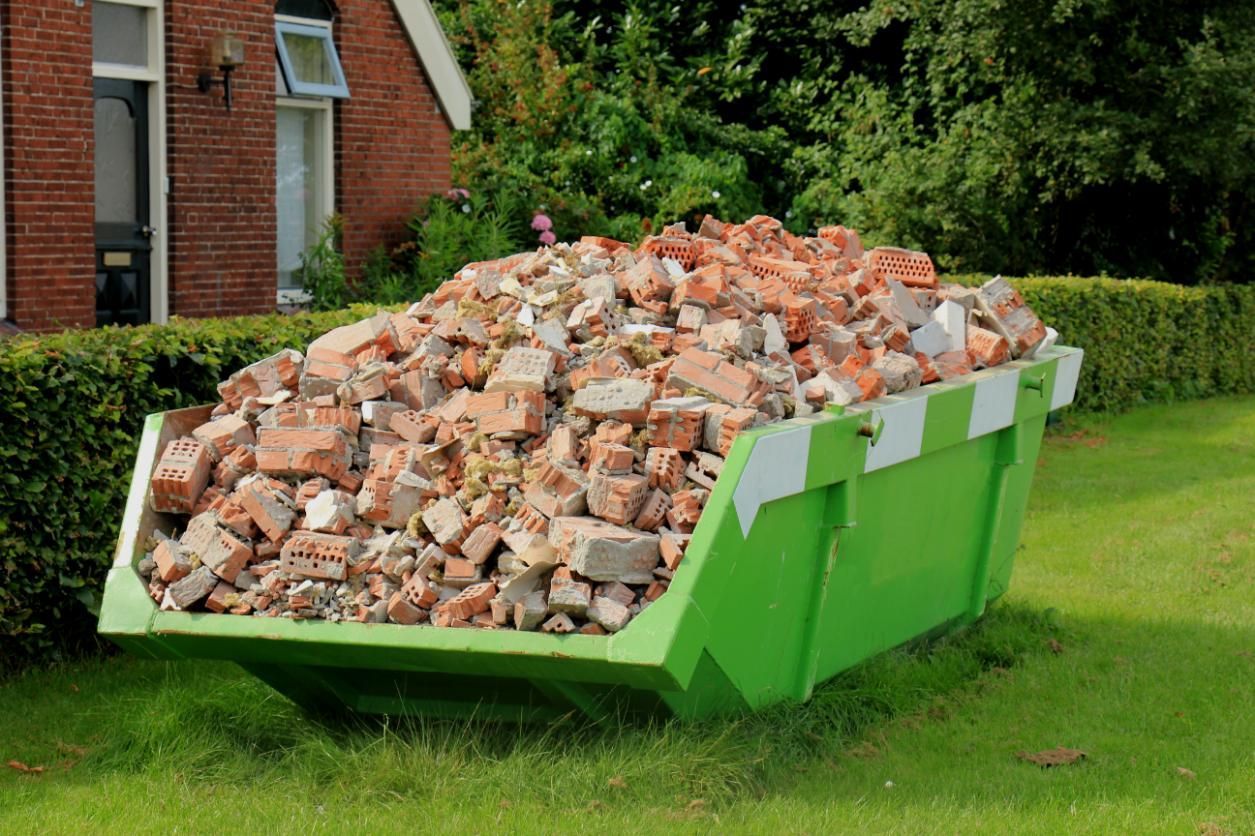 A Green Dumpster Filled With Bricks is Sitting in the Grass in Front of a Brick House — IBS Skip Bins in Cooroy, QLD