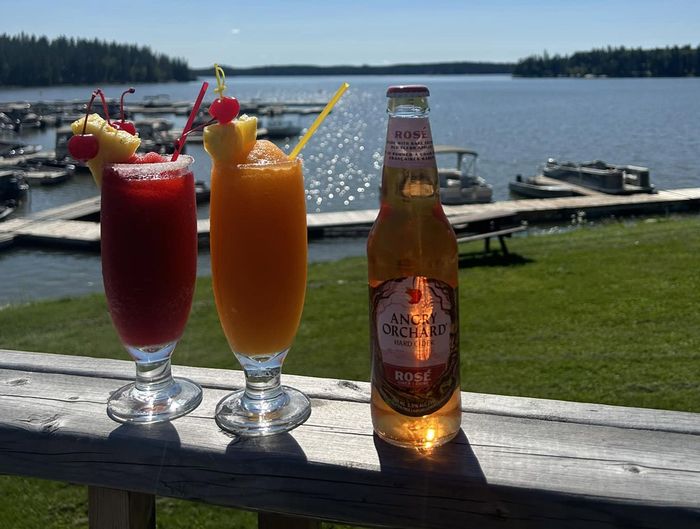 Two drinks and a bottle of beer are on a wooden deck overlooking a lake.