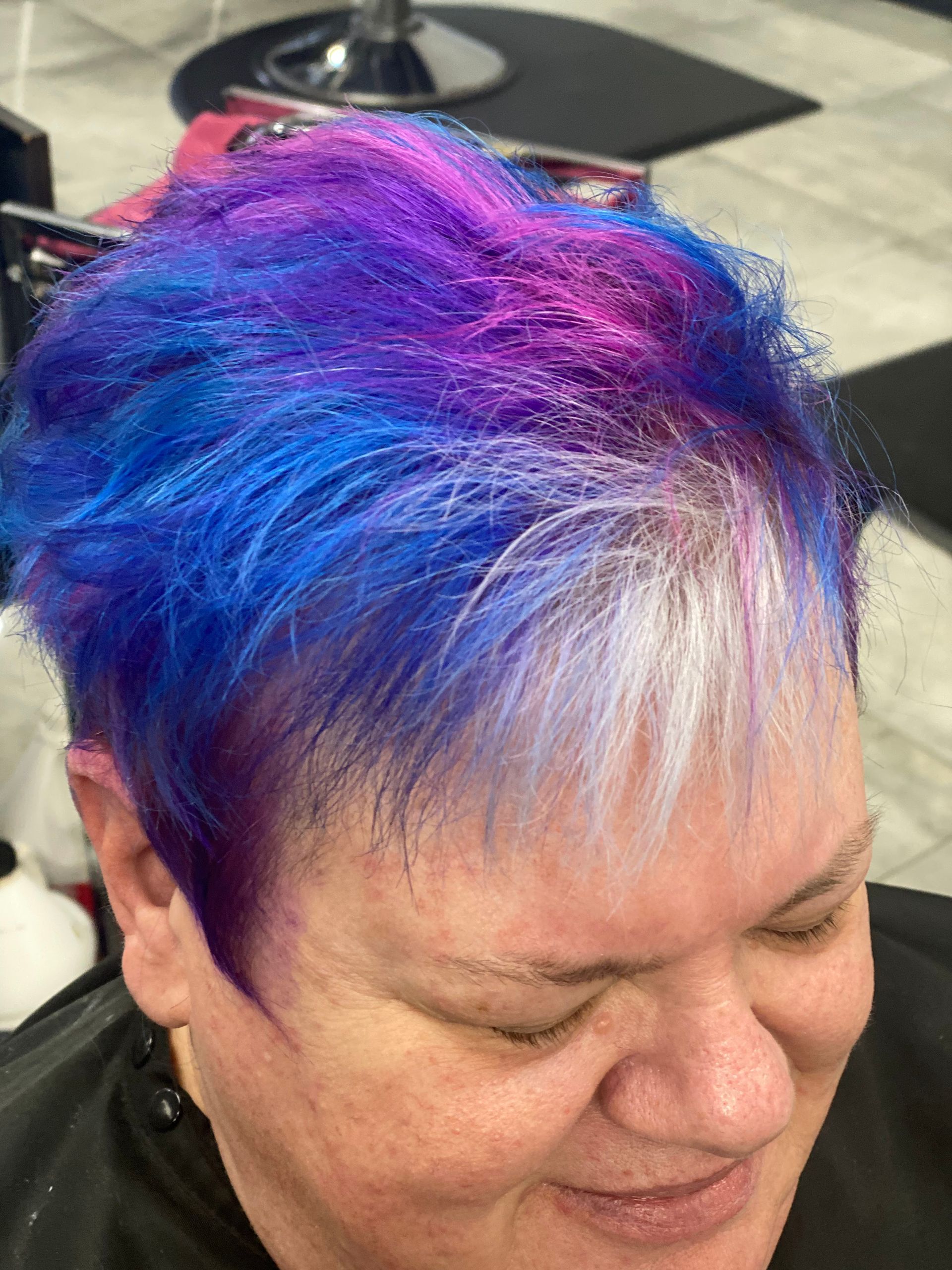 A woman with blue and purple hair is sitting in a chair in a salon.
