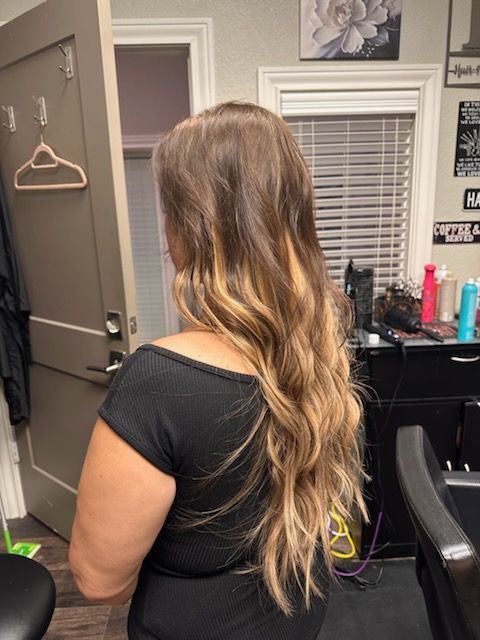 Woman with long wavy blonde hair seated in a salon chair, shown from behind.