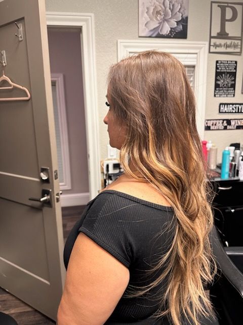Woman with long wavy hair in a salon chair, viewed from the side against a gray wall.