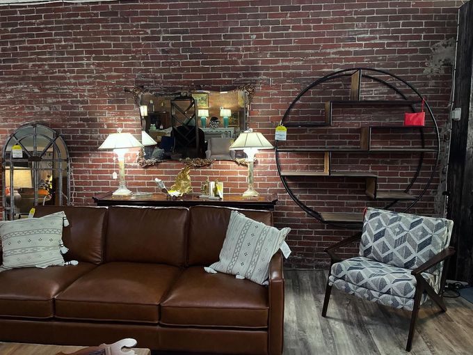 A brown leather sofa, patterned armchair, and circular shelving unit arranged against a rustic red brick wall.