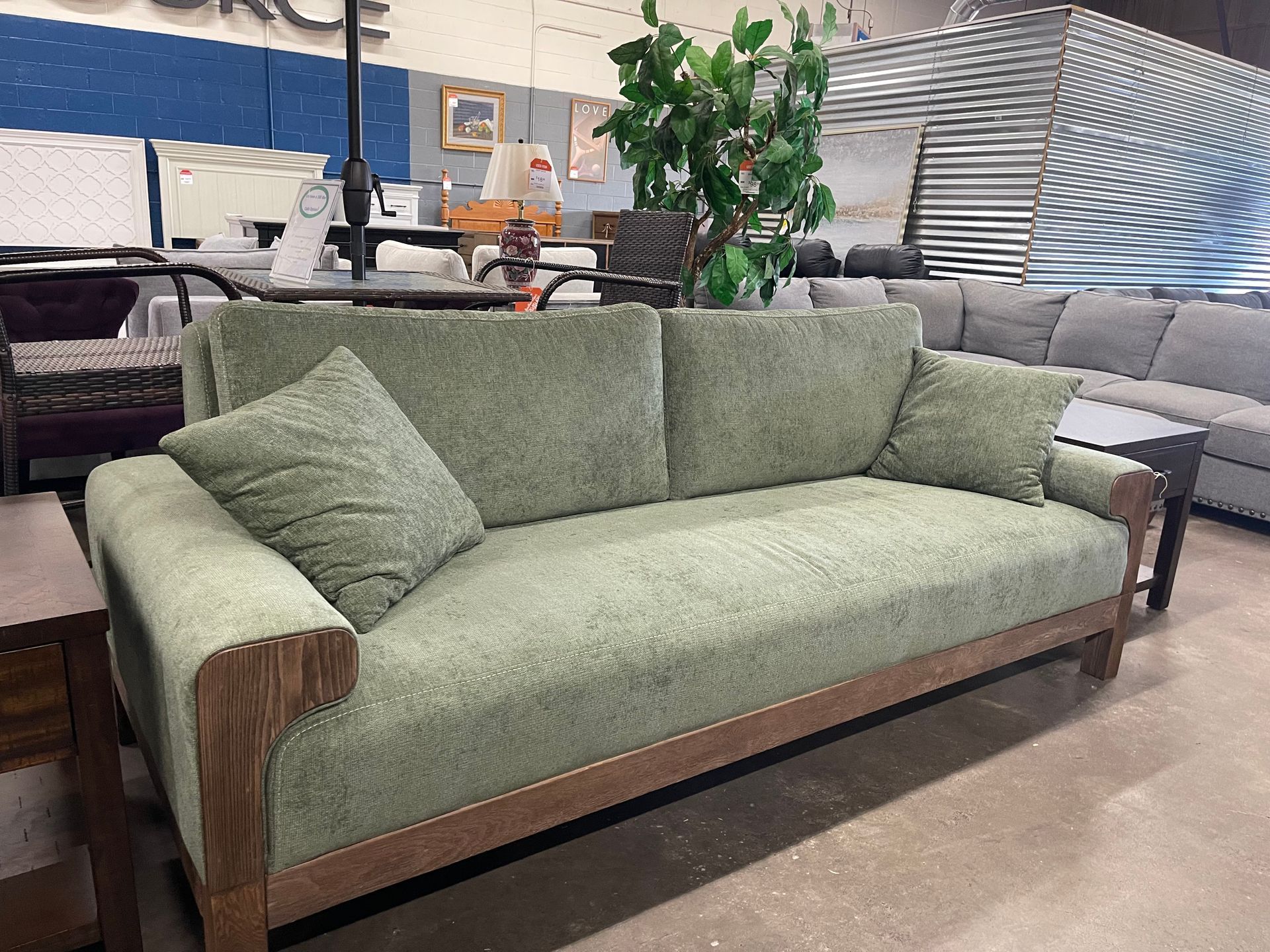 A sage green sofa with a wooden base and two matching throw pillows displayed in a furniture showroom.