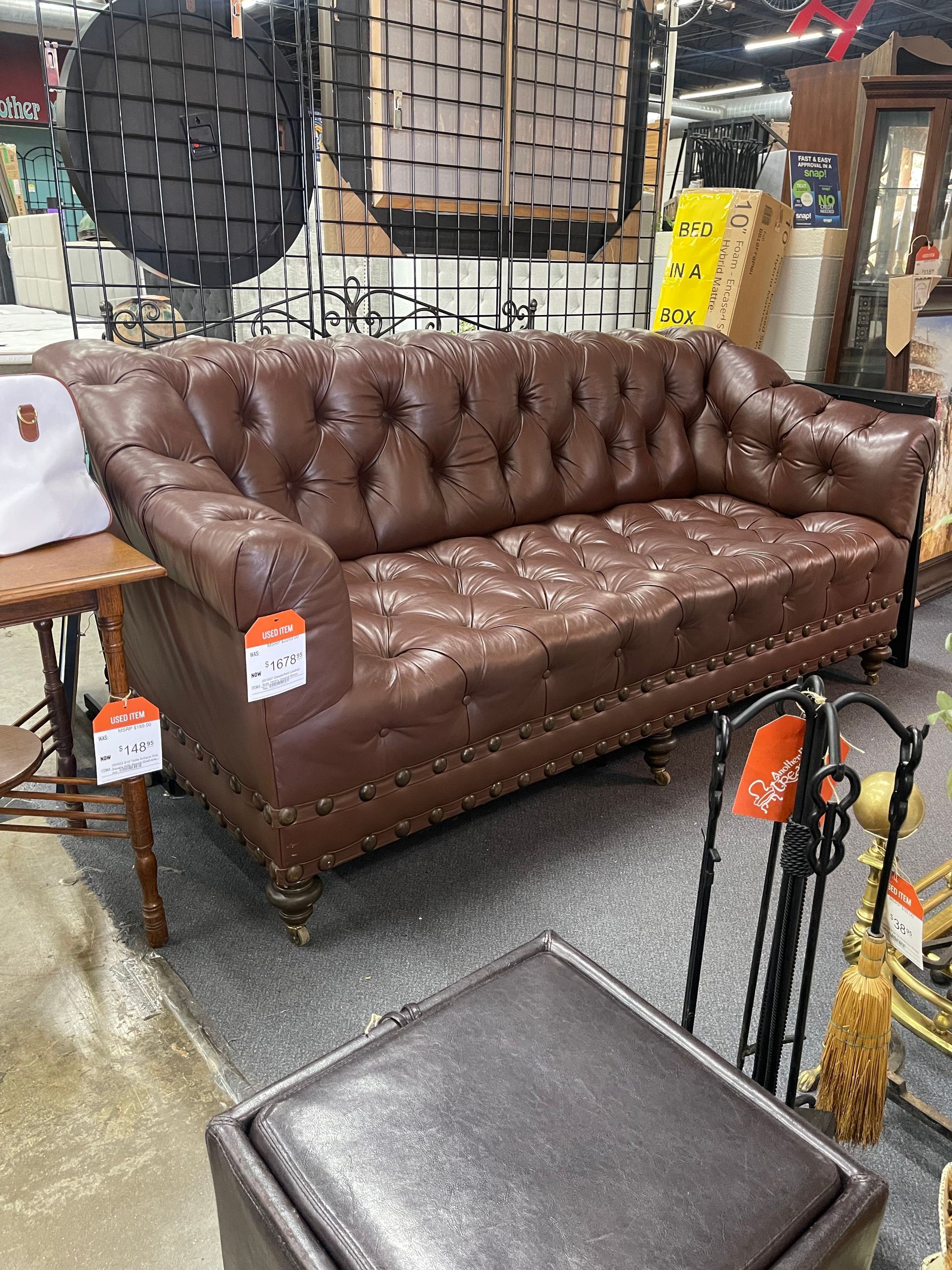 A brown tufted leather sofa with nailhead trim sits in a store, with store tags attached.