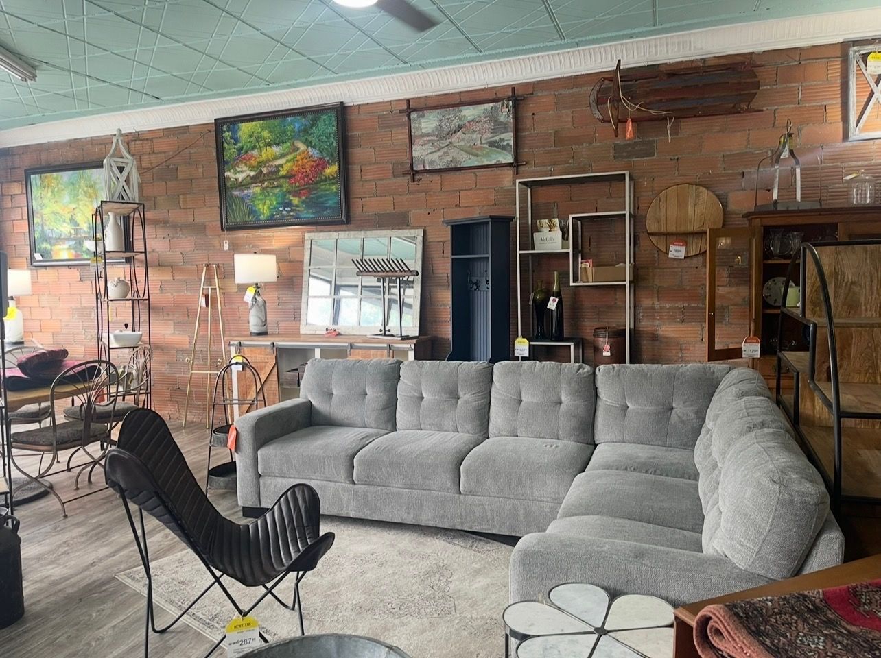 A gray sectional sofa sits in a store showroom against a brick wall decorated with framed art and shelving units.