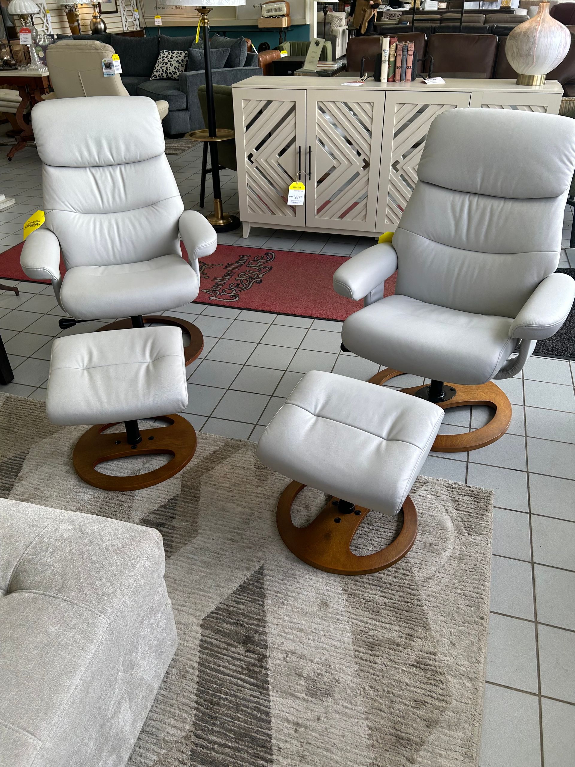 Two matching light gray leather recliners with wooden bases and coordinating ottomans set on a rug in a furniture store.