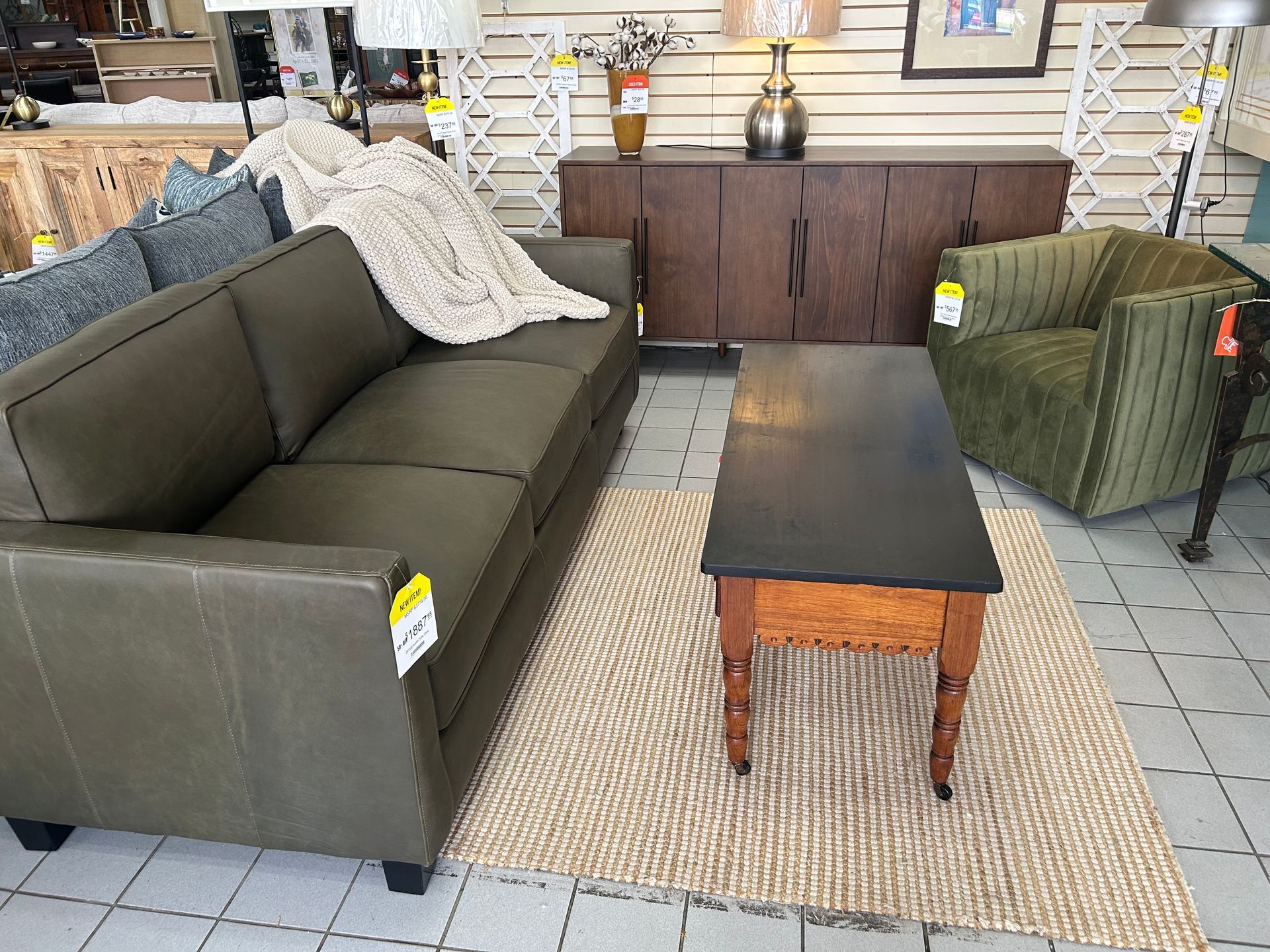 A furniture showroom with an olive green sofa, a dark wooden coffee table, a textured rug, and a green velvet armchair.