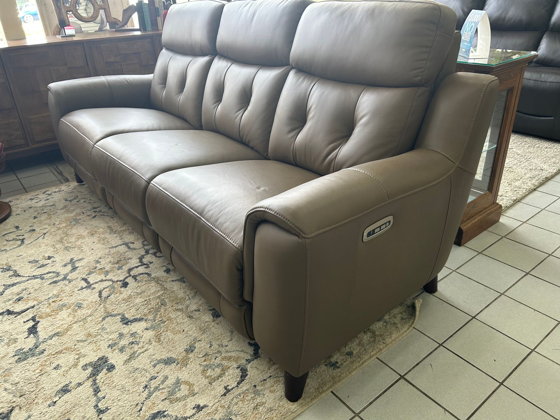 A taupe leather recliner sofa with button-tufted back cushions sits on a patterned area rug in a showroom.