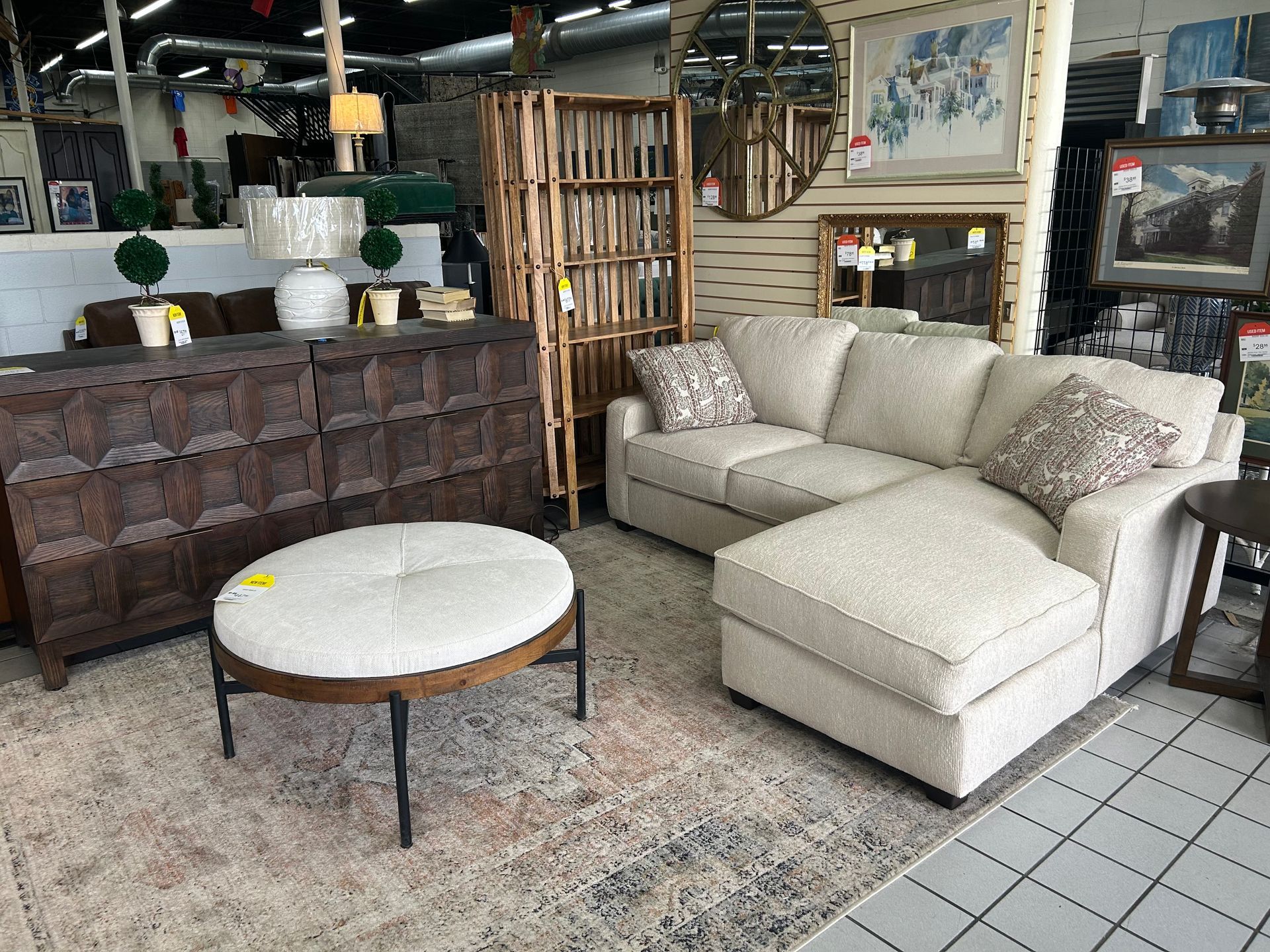 A furniture showroom featuring a beige sectional sofa, a round upholstered coffee table, and a dark wooden credenza.