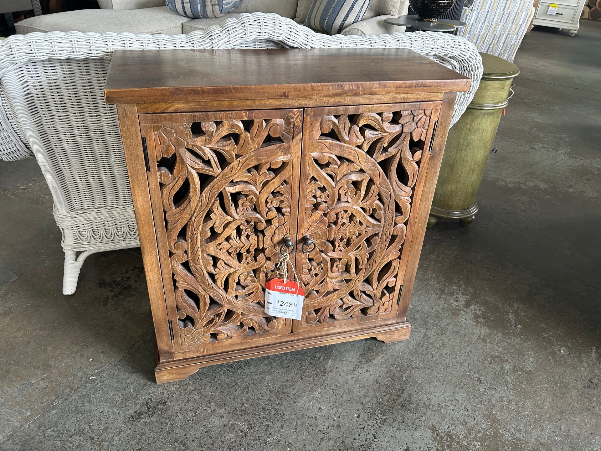 A small, light brown wooden cabinet with intricately carved double doors featuring circular patterns, set in a store.