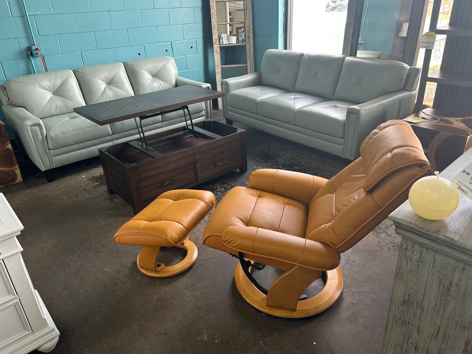 A showroom featuring two light gray sofas, a dark wood lift-top coffee table, and an amber-colored recliner with ottoman.