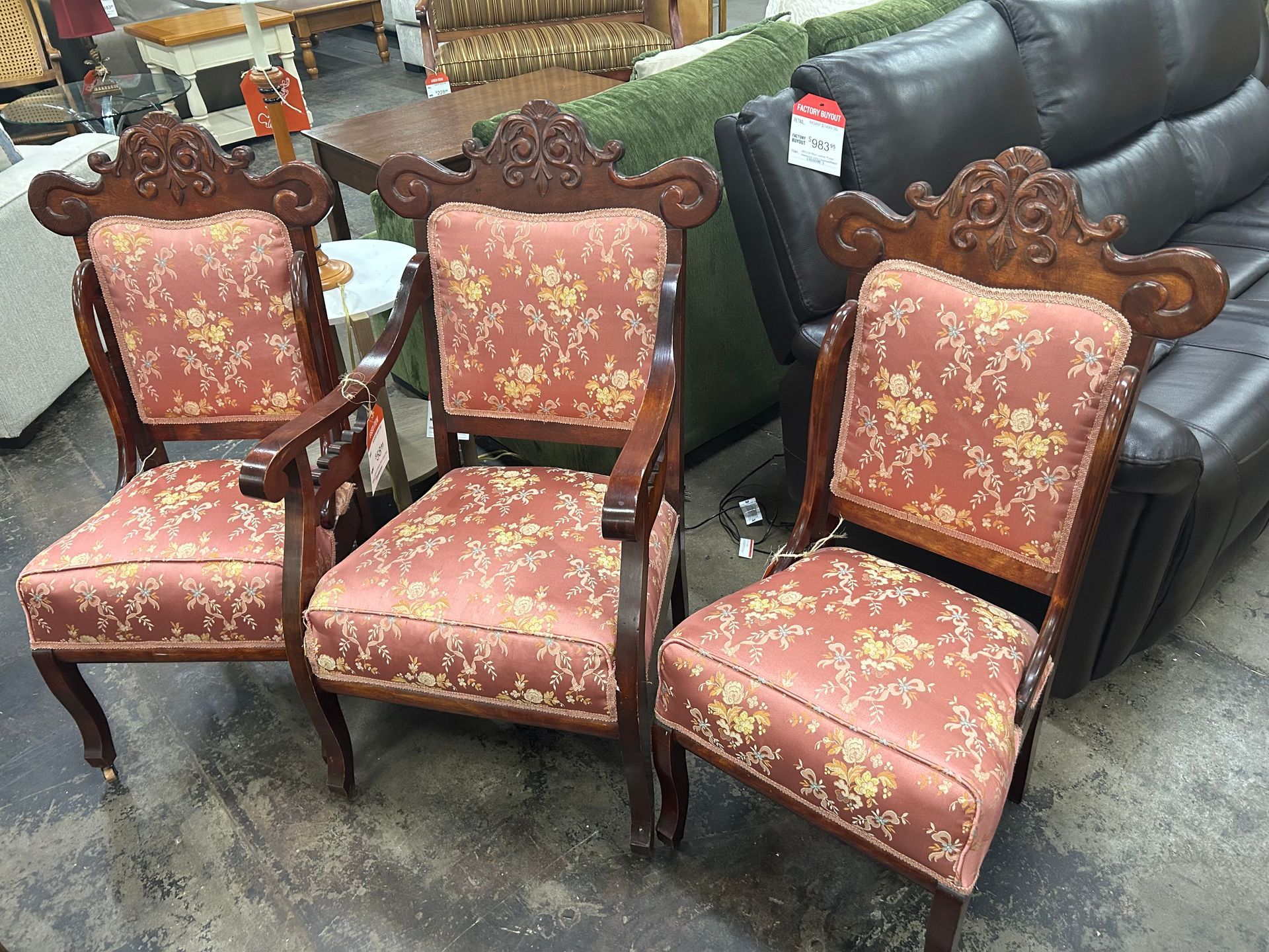Three antique wooden armchairs with pink floral upholstery arranged in a row in a furniture store.
