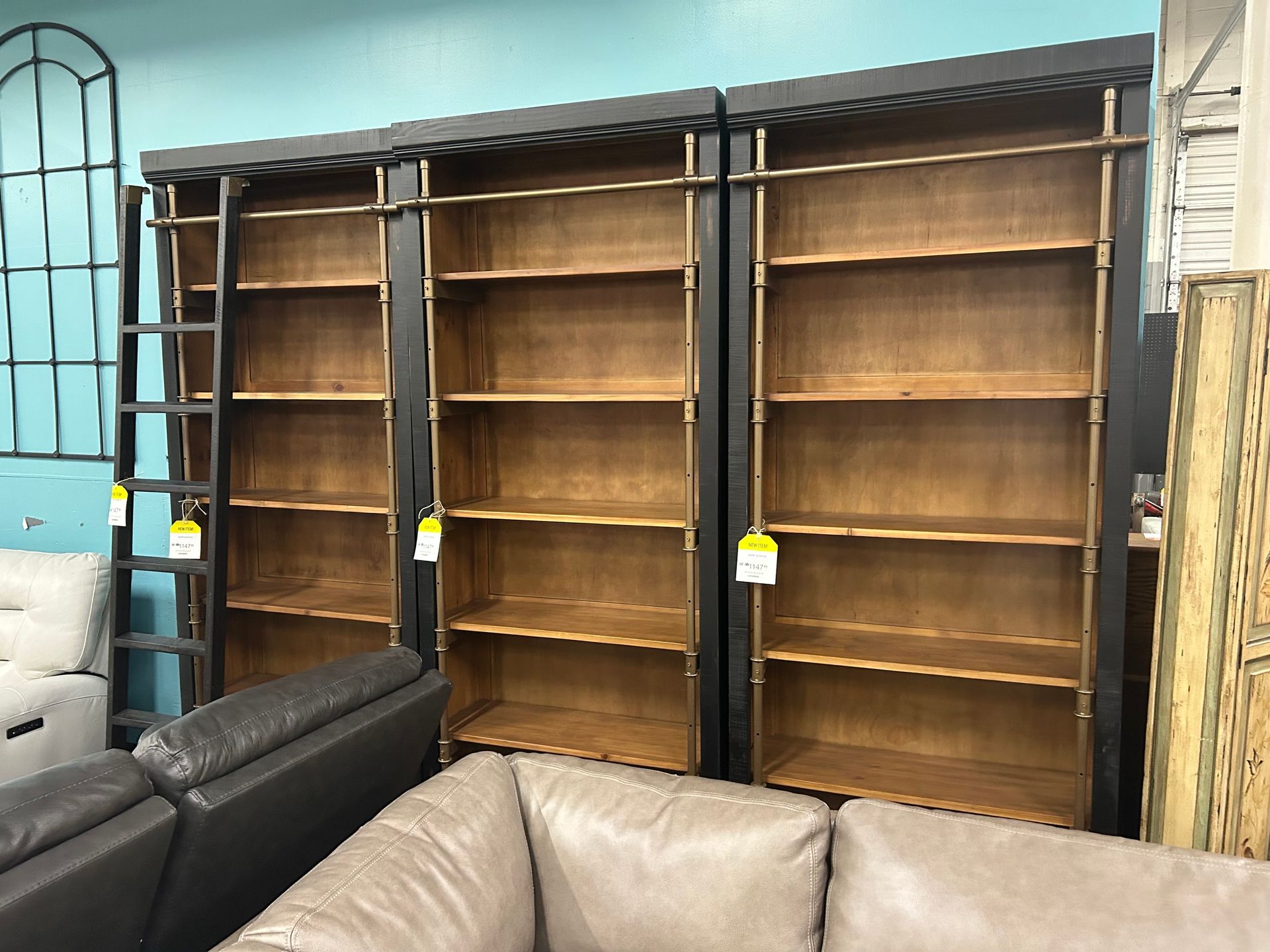 Three tall, dark wood bookshelves with a sliding library ladder in a showroom, positioned behind two leather sofas.