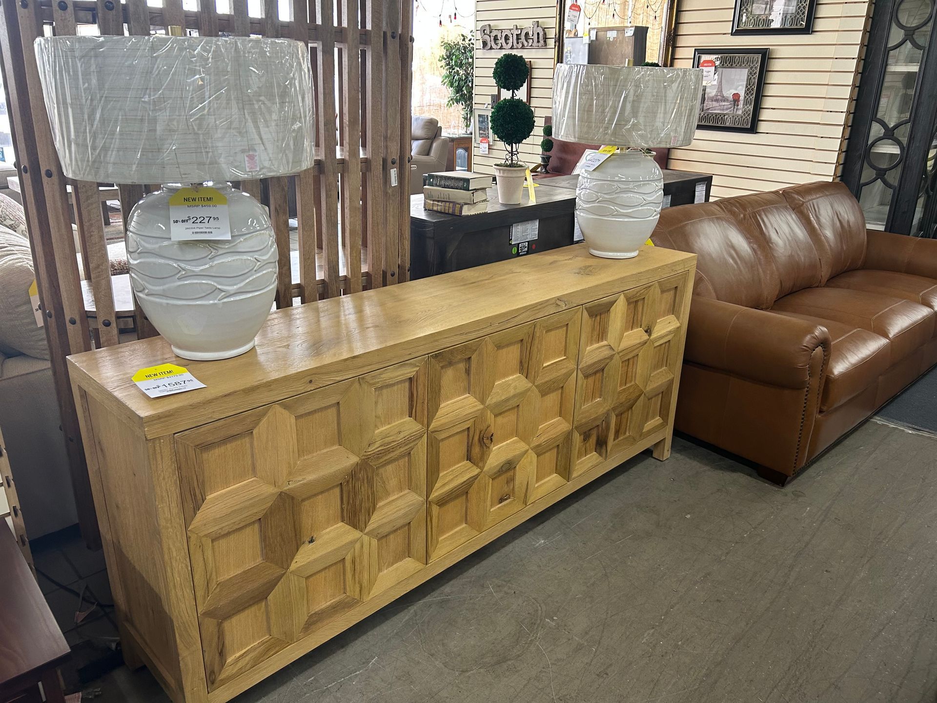 A long, light-wood console table with geometric door panels holds two lamps with white, textured bases in a furniture store.