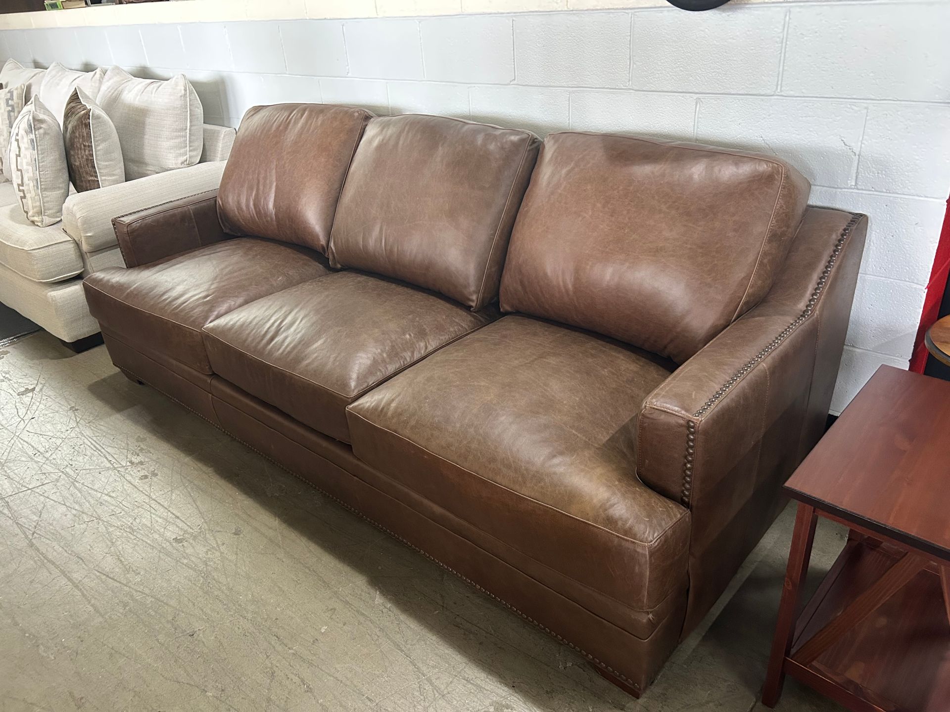 A brown leather three-cushion sofa with stud detailing along the arms, positioned in a room with a white brick wall.