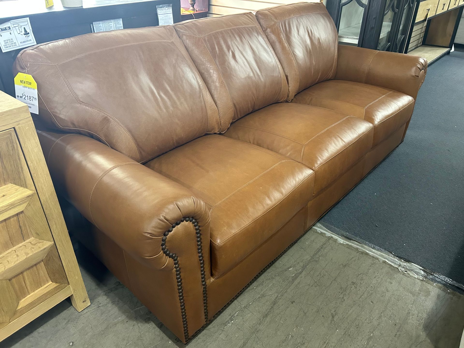 A three-seater brown leather sofa with roll arms and decorative nailhead trim, displayed in a showroom.